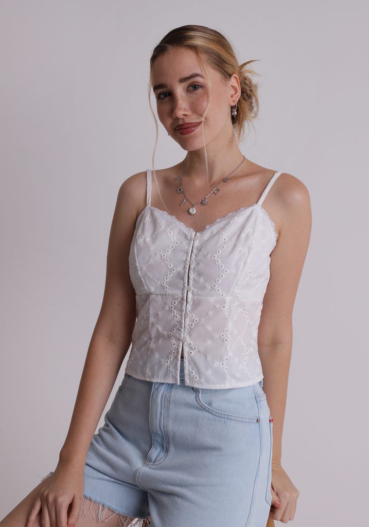 Menina branca, loira, veste blusa branca, com tecido laise, alças finas reguláveis, fechamento frontal por botões e comprimento cropped. Produto com a cor cloud Dancer, a cor de 2026