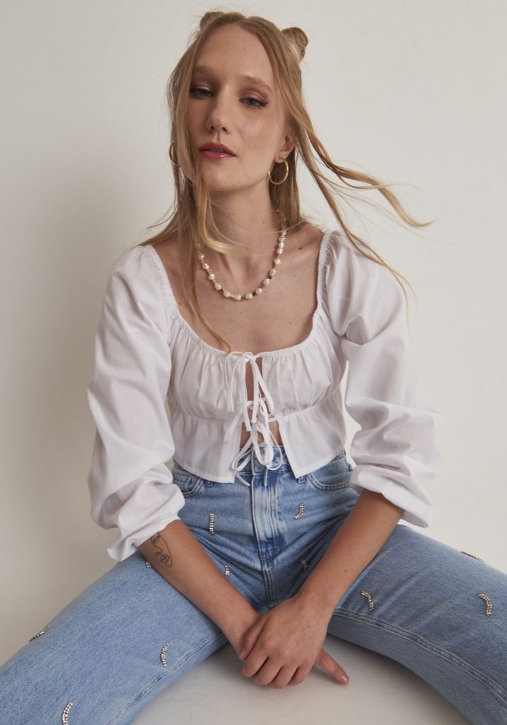 Menina branca, com cabelo semi preso, vestindo blusa estilo bata com, com três amarrações frontais. Produto com a cor cloud Dancer, a cor de 2026